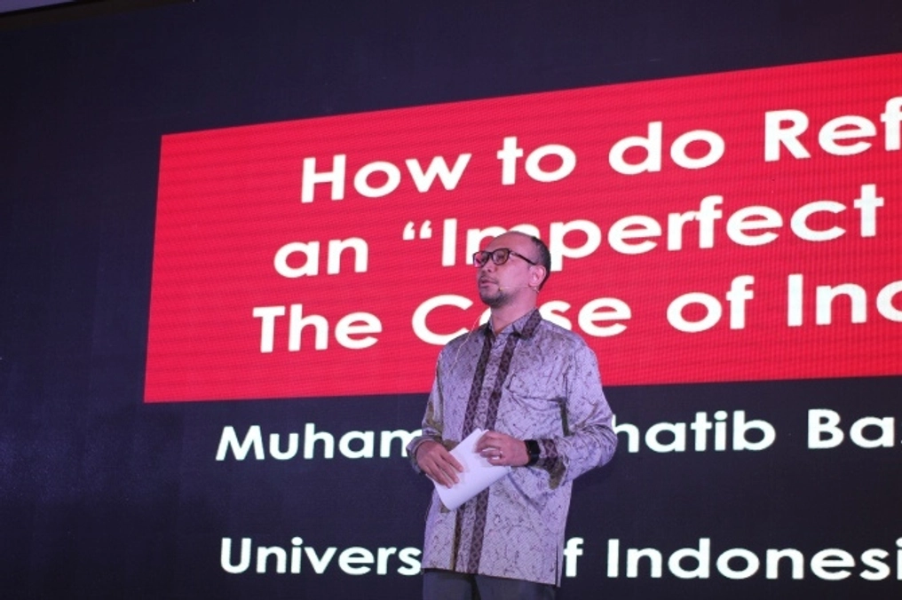 Basri Alf stands on stand with large text on screen behind him.