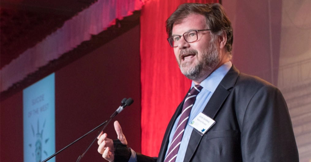 A man with glasses and a beard speaks at a podium.